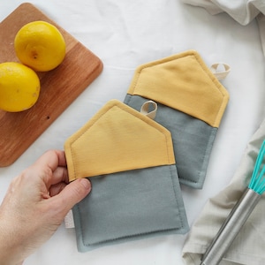 Può includere: Due presine grigie e gialle a forma di casa. Le presine sono fatte di tessuto e hanno un anello per appenderle.