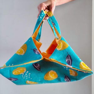 Può includere: Una borsa tote turchese con interno e manici gialli. La borsa presenta un motivo di limoni gialli e foglie viola scuro. La borsa è tenuta da una mano.