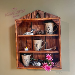 May include: A rustic, wooden shelf with three tiers, stained a warm brown. It features a cutout tree design and holds decorative items like mugs with deer and moose designs, a loon figurine, a pinecone, a candle, and pink flowers. The text "RUSTICONTHERIDGE etsy.com" is visible.