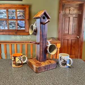 May include: A rustic wooden mug holder in a birdhouse design, stained in shades of brown. It holds four mugs, with three mugs hanging on the holder. The holder is on a wooden base, and the text "Holds 4 mugs" is visible.