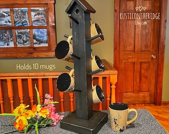 May include: Black wooden mug rack shaped like a birdhouse with a birdhouse roof. The rack holds 7 mugs and has a sign that says "Rusticontheridge Etsy.com". The rack is on a black countertop with a bouquet of yellow and pink flowers.