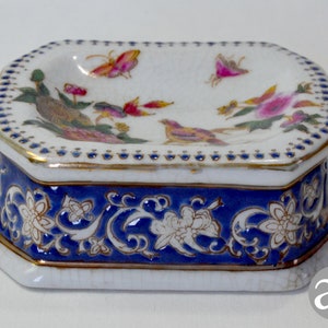 May include: An octagonal porcelain trinket box with a white lid and a blue base. The lid is decorated with painted birds, butterflies, and flowers. The base has a blue floral design with gold accents. A decorative item.