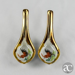 Pode incluir: Duas colheres de cerâmica douradas com interior branco e design de galo. O galo é retratado em tons de vermelho, preto e laranja, com detalhes de folhagem verde. As colheres têm um acabamento brilhante e são provavelmente para servir.