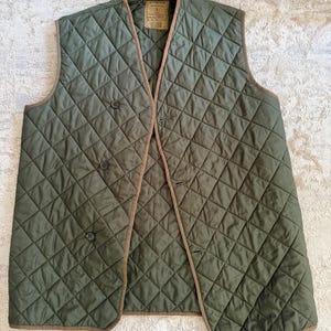 May include: Olive green quilted vest with a diamond pattern and brown trim. The vest has a button-up front and a label on the inside. A classic outerwear piece.