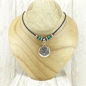 Peut inclure: Un collier pendentif argenté avec un pendentif circulaire orné d'un motif animalier détaillé. Le collier est composé d'un cordon noir avec des perles turquoise et crème. Le pendentif est présenté sur un buste à bijoux en bois.
