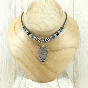 May include: A silver pendant necklace with a black cord. The necklace features an arrow-shaped pendant, and is accented with black, turquoise, and zebra-striped beads. The necklace is displayed on a wooden bust.