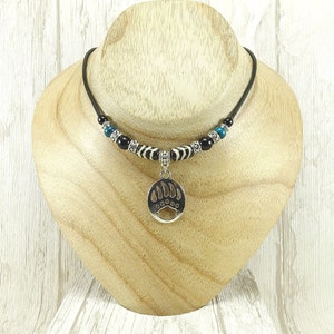 May include: A black cord necklace with a silver bear paw pendant. The necklace has black, turquoise, and zebra-striped beads. The pendant is oval-shaped with a bear paw print. The necklace is displayed on a wooden bust.