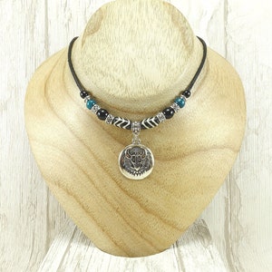 Peut inclure: Un collier en cordon noir avec un pendentif argenté représentant un taureau. Le collier comporte des perles turquoise, noires et à motifs. Le pendentif est rond et présente une image détaillée d'une tête de taureau.