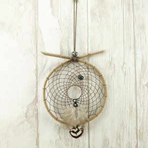 May include: A handmade dreamcatcher with a circular woven web, decorated with feathers and beads. The dreamcatcher is suspended by a brown cord and has a wooden frame. The background is a light-coloured wooden wall.