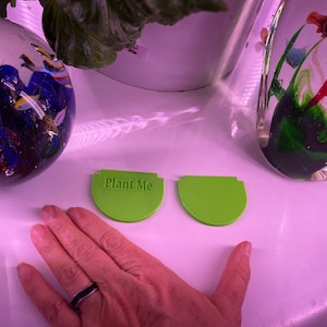 May include: Two green plastic plant markers, one with the text "Plant Me" on it. The markers are on a white surface.