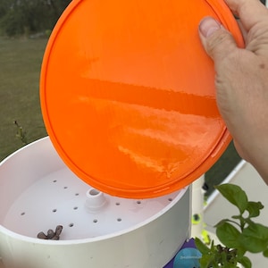 May include: A white plastic hydroponic gardening system with an orange lid. The lid is being lifted off to reveal the system's interior. The system has a purple label with the text "ZHUGONGMANGGU" on it.