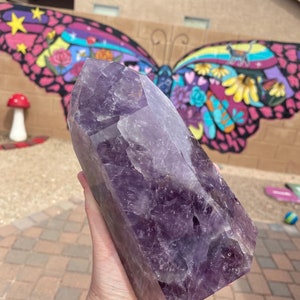 May include: A large, deep purple amethyst crystal point with a natural, faceted shape. The crystal is held in a hand, showcasing its size and intricate details.