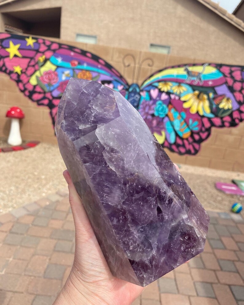 May include: A large, deep purple amethyst crystal point with a natural, faceted shape. The crystal is held in a hand, showcasing its size and intricate details.