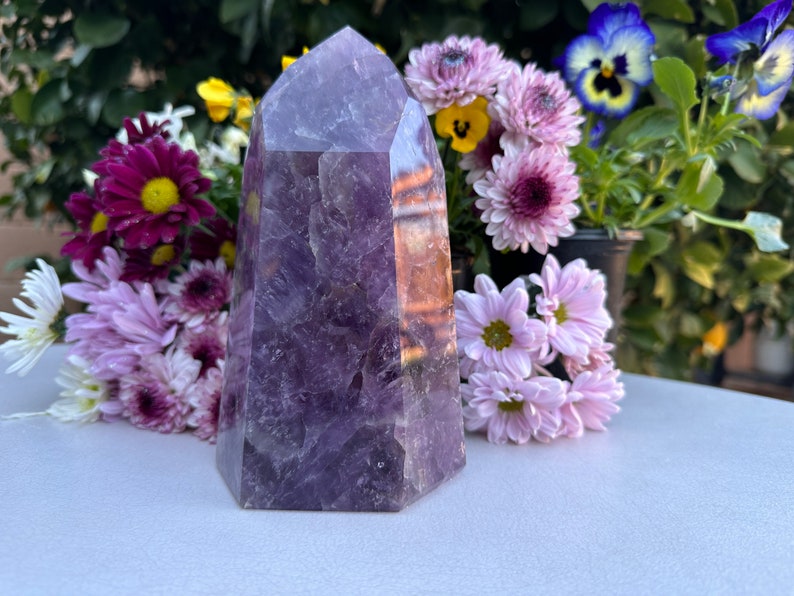 May include: A large, purple amethyst crystal point with a clear, faceted top. The crystal is sitting on a white surface with a bouquet of flowers behind it.