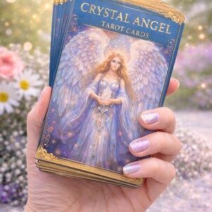 Op de afbeelding: Een hand houdt een pakje "Crystal Angel Tarot Cards" vast. Het kaartontwerp toont een hemelse engel in een vloeiende jurk, die een kristal vasthoudt. De kaarten hebben een blauwe achtergrond met gouden accenten en de tekst "Crystal Angel Tarot Cards".