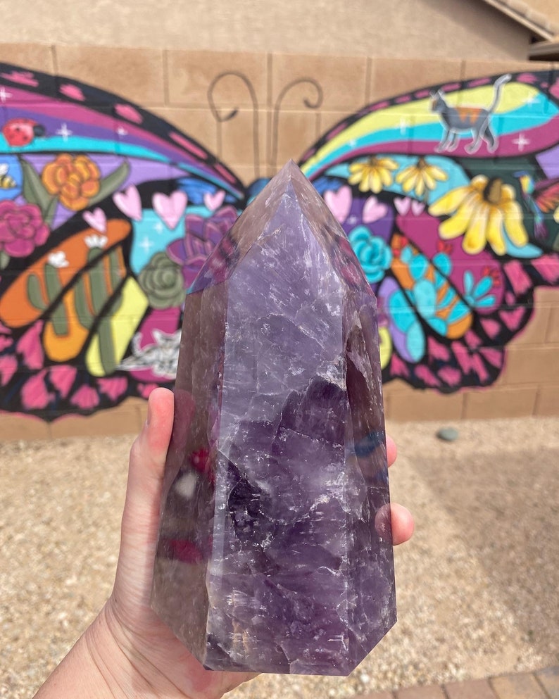 May include: A large, purple amethyst crystal point with a natural, faceted shape. The crystal is held in a hand, with a colorful butterfly mural in the background.