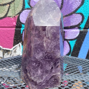 May include: A large, purple amethyst crystal point with a clear, faceted top. The crystal is sitting on a metal surface.