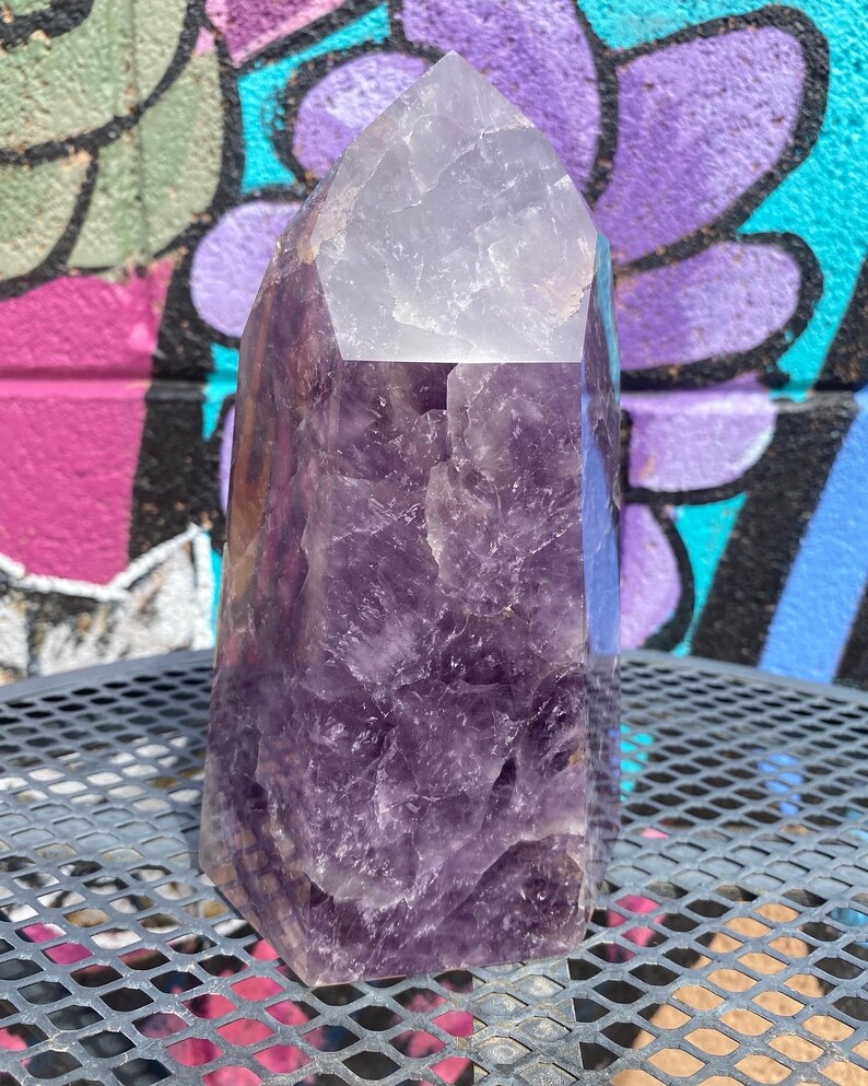 May include: A large, purple amethyst crystal point with a clear, faceted top. The crystal is sitting on a metal surface.