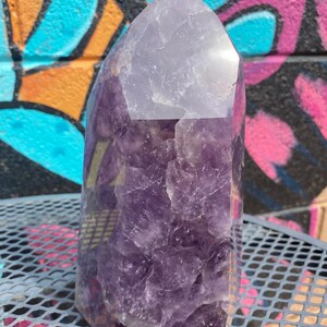 May include: A large, purple amethyst crystal point with a clear, faceted top. The crystal has a natural, rough texture and is a beautiful example of this popular gemstone.