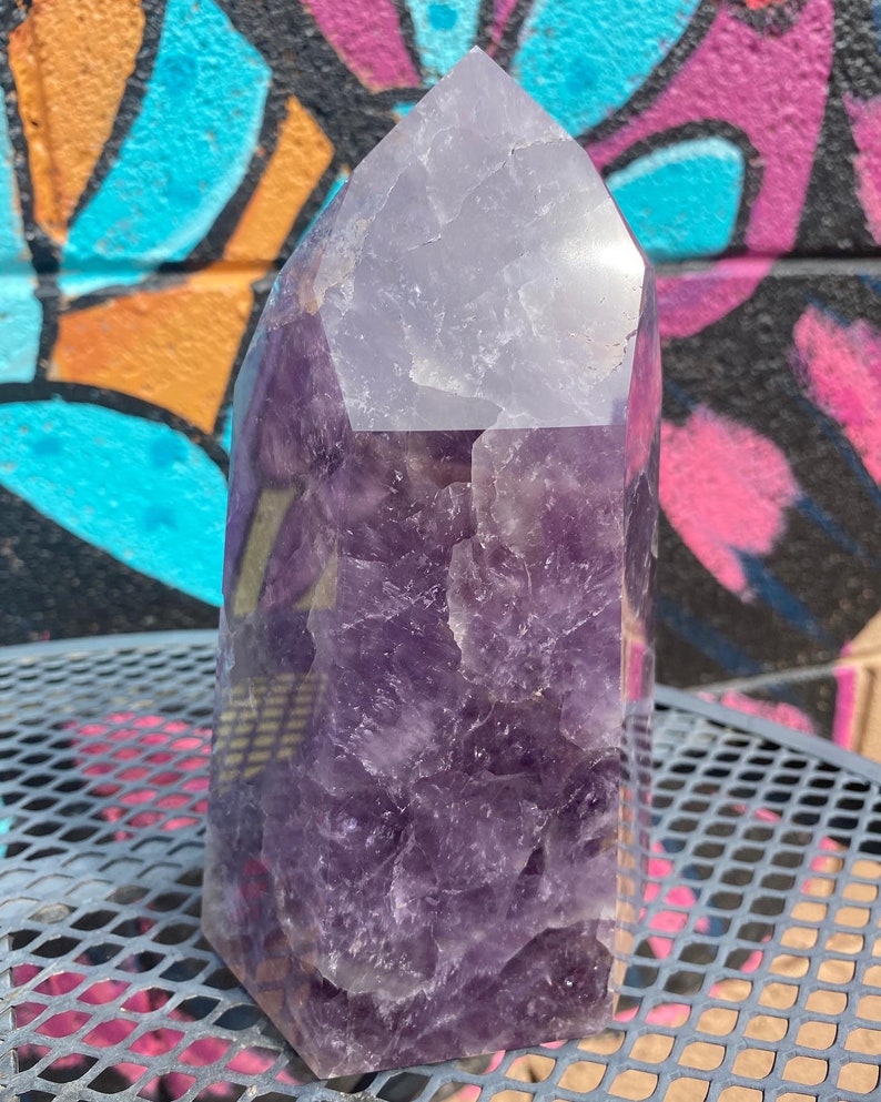 May include: A large, purple amethyst crystal point with a clear, faceted top. The crystal has a natural, rough texture and is a beautiful example of this popular gemstone.