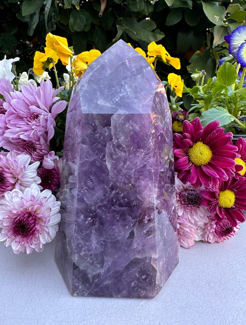 May include: A large, purple amethyst crystal point with a faceted, geometric shape. The crystal is surrounded by a variety of flowers, including pink and yellow blossoms.