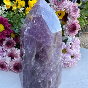May include: A large, purple amethyst crystal point with a faceted, geometric shape. The crystal is sitting on a light gray surface, surrounded by purple and yellow flowers.
