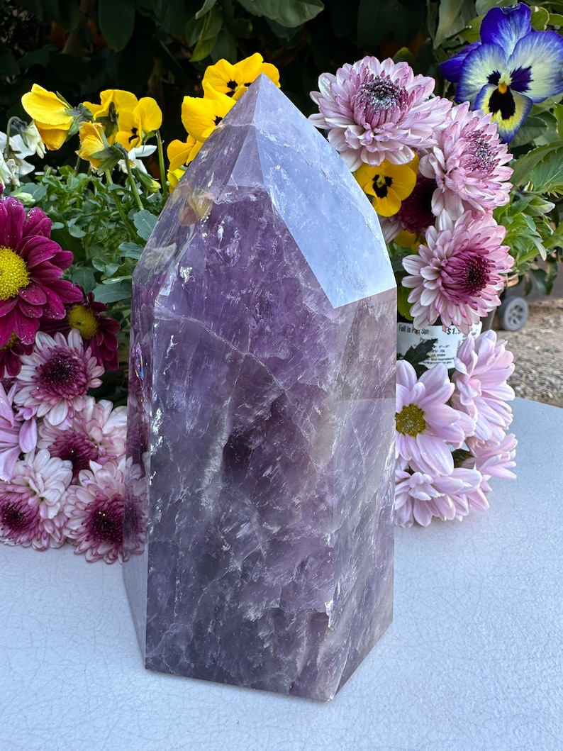May include: A large, purple amethyst crystal point with a faceted, geometric shape. The crystal is sitting on a light gray surface, surrounded by purple and yellow flowers.