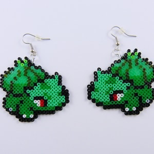 以下が含まれることがあります： ポケモンのキャラクターの形をした緑と黒のビーズのイヤリングのペア。イヤリングは銀色のフックにぶら下がっています。