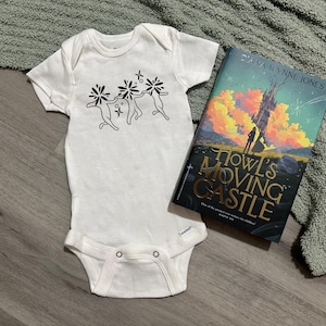 Puede incluir: Body de beb&eacute; blanco con un gr&aacute;fico negro de tres figuras tomadas de la mano con detalles florales. Junto a &eacute;l, un ejemplar de "El castillo ambulante" de Diana Wynne Jones, con una ilustraci&oacute;n de castillo en la portada.