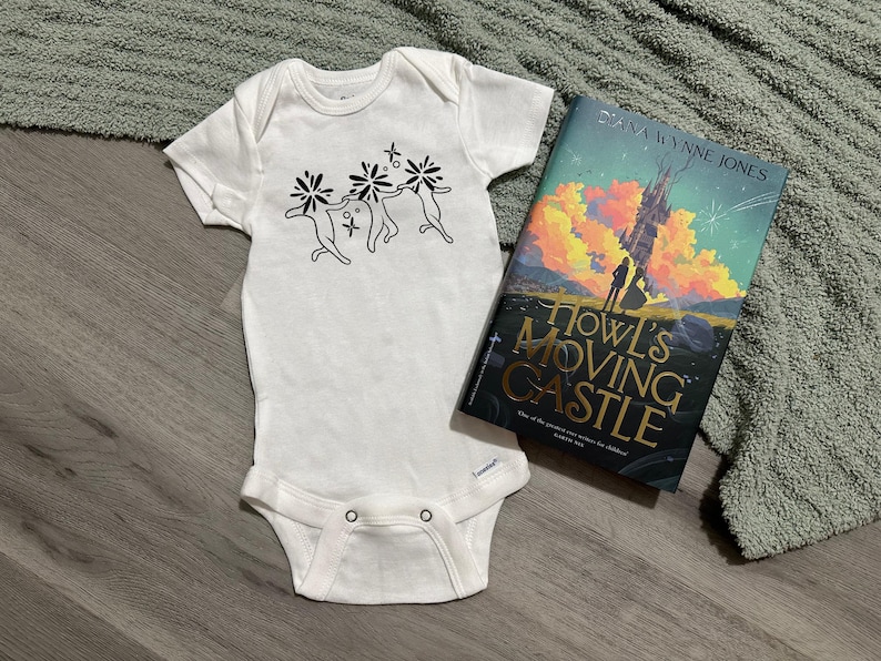 Puede incluir: Body de beb&eacute; blanco con un gr&aacute;fico negro de tres figuras tomadas de la mano con detalles florales. Junto a &eacute;l, un ejemplar de "El castillo ambulante" de Diana Wynne Jones, con una ilustraci&oacute;n de castillo en la portada.