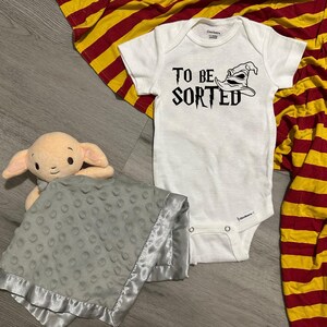 May include: A white baby onesie with the text "TO BE SORTED" and a Sorting Hat graphic. A plush doll and a gray blanket with a satin trim are also in the image. A red and yellow striped blanket is in the background.