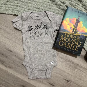 Puede incluir: Un body de beb&eacute; gris con un gr&aacute;fico negro de tres figuras estilizadas con cabezas florales. Junto a &eacute;l, un ejemplar de "El castillo ambulante" de Diana Wynne Jones, con una colorida ilustraci&oacute;n de portada.