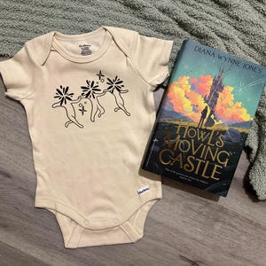 Puede incluir: Un body de beb&eacute; color crema con un gr&aacute;fico negro de tres figuras bailando con acentos florales. Junto al body hay un ejemplar de "El castillo ambulante" de Diana Wynne Jones.