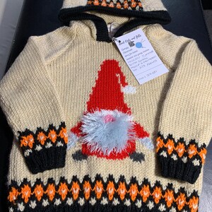 May include: A cream knitted hooded sweater with a red and white gnome design. The sweater has a black and orange trim and a matching hood. The tag reads "Hand Knits and Gifts" and "Hooded Jumper with Grey  Mohair & a Pattern for 2-3 Years Price: £29.00"