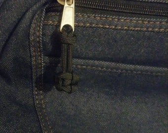 Puede incluir: Primer plano de un tirador de cremallera en una bolsa de mezclilla. El tirador de cremallera está hecho de paracord negro y tiene un nudo en el extremo. La cremallera es plateada y tiene una cinta de cremallera negra.
