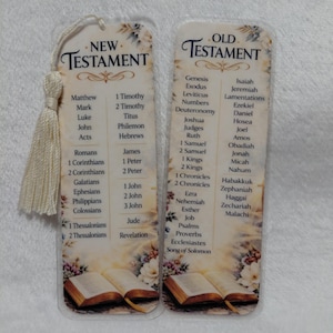 Könnte beinhalten: Zwei transparente Acryl-Lesezeichen mit Quaste, die Bücher des Alten und Neuen Testaments auflisten. Die Lesezeichen haben florale Akzente und Bilder von offenen Büchern unten. Der Text ist schwarz.