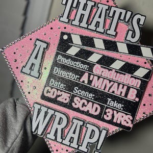 Könnte beinhalten: Eine rosa Abschlusskappe mit den Worten "THAT'S A WRAP!" in weißer und silberner Schrift. Ein schwarz-silbernes Klapptafel-Design enthält den Text "Graduation", "A'MIYAH B.", "CO'25", "SCAD" und "3 YRS."