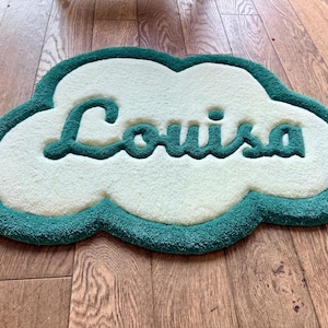 May include: A cloud-shaped tufted rug featuring a cream-coloured centre and a dark teal border. The name Louisa is written in the centre in a dark teal cursive script. The rug has a thick, textured pile and a small white tag on the left side.