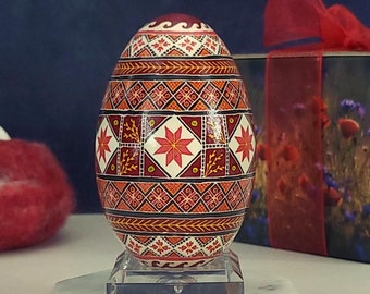 May include: A hand-painted Easter egg with a geometric pattern in shades of red, orange, and white. The egg is displayed on a white pedestal.