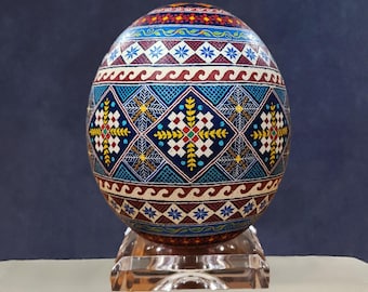May include: A hand-painted Easter egg with intricate geometric patterns in shades of blue, red, yellow, and white. The egg is displayed on a clear acrylic stand.