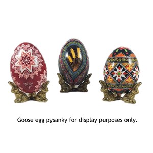 May include: Three intricately decorated goose eggs, each with a unique design, are displayed on gold metal bunny-shaped stands. The eggs are painted in vibrant colors, including red, blue, black, white, and yellow. The text "Goose egg pysanky for display purposes only." is visible below the eggs.