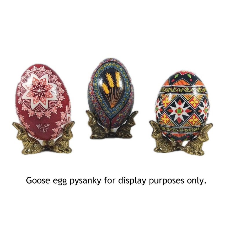 May include: Three intricately decorated goose eggs, each with a unique design, are displayed on gold metal bunny-shaped stands. The eggs are painted in vibrant colors, including red, blue, black, white, and yellow. The text "Goose egg pysanky for display purposes only." is visible below the eggs.