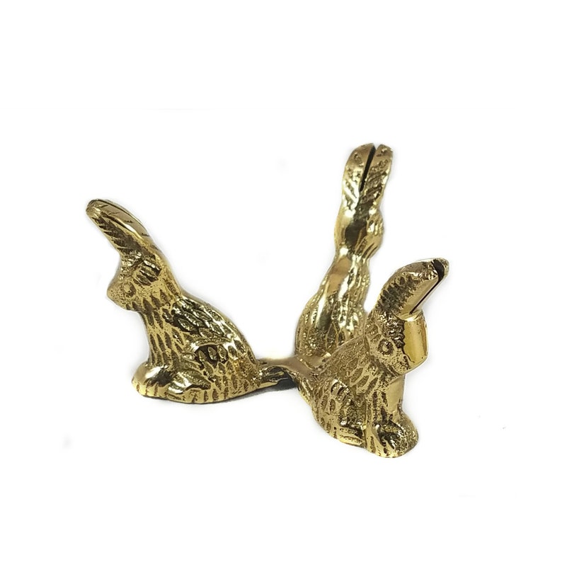 May include: A gold metal egg holder shaped like two rabbits. The rabbits are facing each other with their heads up and their tails down. The holder is made of a smooth, shiny metal.