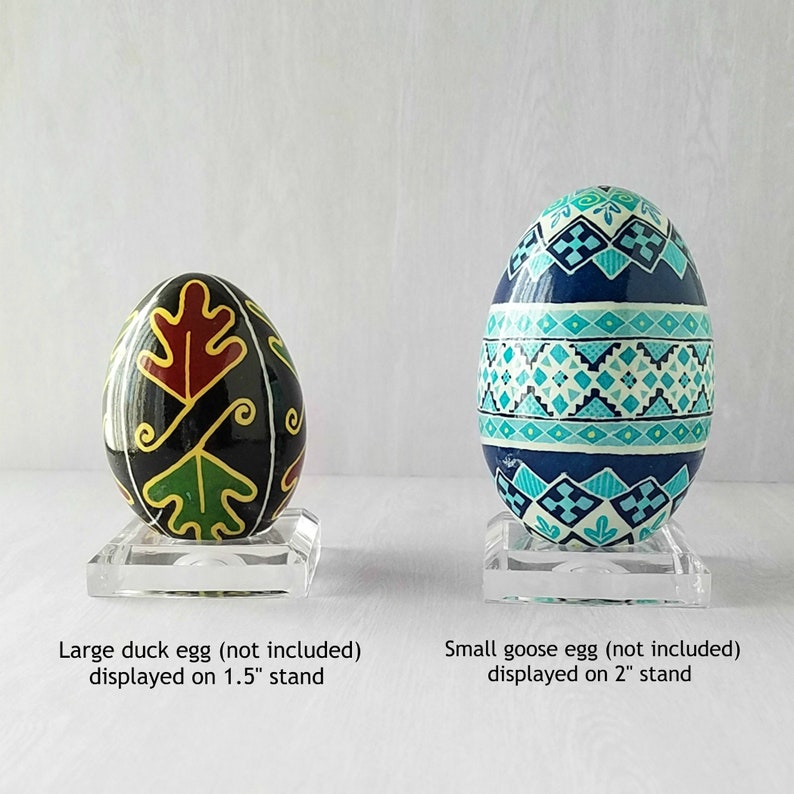 May include: Two hand-painted Easter eggs on clear acrylic stands. The larger egg is black with green and red leaf designs and is on a 1.5 inch stand. The smaller egg is blue with white and turquoise geometric designs and is on a 2 inch stand.
