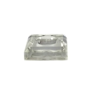 May include: A clear glass display stand with a square base and a circular indentation in the center. The stand is designed to hold small objects, such as figurines or jewelry.