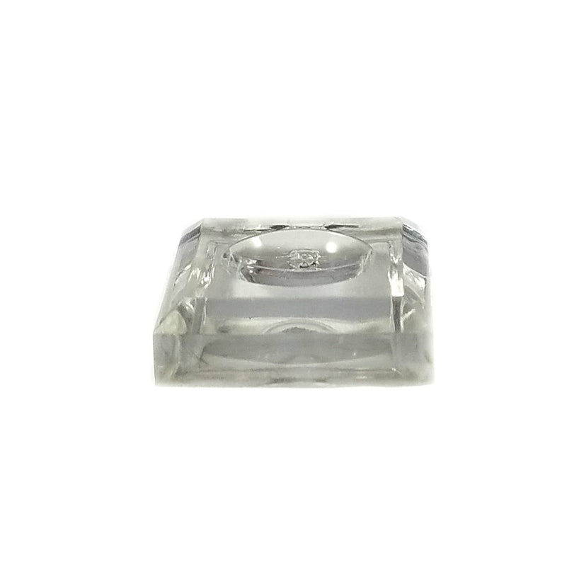 May include: A clear glass display stand with a square base and a circular indentation in the center. The stand is designed to hold small objects, such as figurines or jewelry.