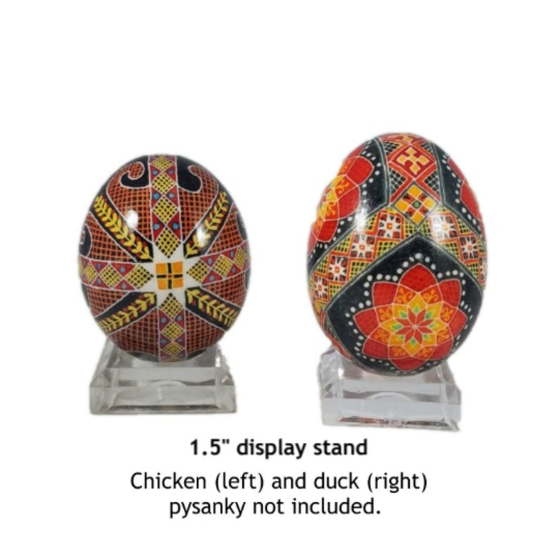 May include: Two 1.5 inch clear acrylic display stands for pysanky eggs. The stands are 1.5 inches tall and have a round base. The stands are shown with two decorated eggs, one with a red, yellow, and black design and the other with a red, orange, and black design.  The text "1.5" display stand" is below the stands. The text "Chicken (left) and duck (right) pysanky not included." is below the text "1.5" display stand".