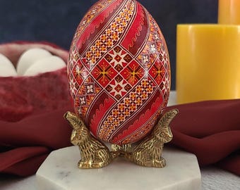 May include: A patterned Easter egg in red, white, and yellow, displayed on a gold-coloured bunny stand. The egg features a detailed geometric design. The stand sits on a white marble base. A basket of white eggs is in the background.