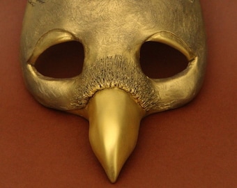 May include: A gold-colored mask with a beak-like nose and feathery details around the eyes. The mask is made of a smooth, textured material.