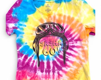 Puede incluir: Una camiseta de manga corta con un vibrante patrón tie-dye en tonos azul, amarillo, rosa y naranja. El gráfico central muestra una ilustración negra de gafas de aviador y un gorro con el texto SASHA GO! impreso en letras negritas.